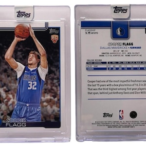 May include: A Topps basketball trading card featuring Cooper Flagg of the Dallas Mavericks. The front shows Flagg in a blue jersey shooting a basketball. The back includes player stats and biographical information. The card is encased in a protective holder.