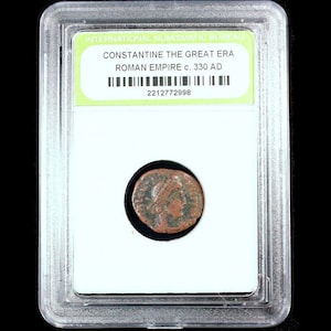 Op de afbeelding: Een Romeinse munt uit de tijd van Constantijn de Grote, ca. 330 na Christus, in een beschermende houder. De munt is bronskleurig met een profiel van een persoon. De houder heeft een groen en wit label met tekst.