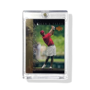 Puede incluir: Un porta tarjetas de plástico transparente con una tarjeta dorada en su interior. La tarjeta presenta una foto de un golfista balanceando un palo. El texto de la tarjeta dice "Tiger's Tales" y "Upper Deck." La tarjeta también incluye una firma que dice "Tiger Woods." El texto en la parte inferior de la tarjeta dice "Tiger goes to Stanford, 1994-96."