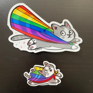 Könnte beinhalten: Zwei Aufkleber mit einer grauen Katze, die einen Regenbogen-Umhang trägt. Der erste Aufkleber zeigt die Katze, die mit einem Regenbogen hinter sich durch die Luft fliegt. Der zweite Aufkleber zeigt die Katze, die mit ausgestreckten Pfoten fliegt. Beide Aufkleber haben den Text "stephnderek.com" auf sich.