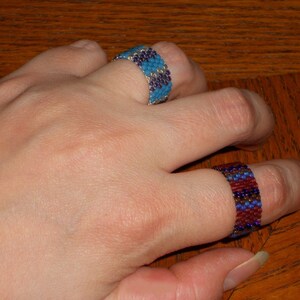 May include: Two beaded rings on a hand. The top ring is blue and silver, the bottom ring is red, blue, and silver.