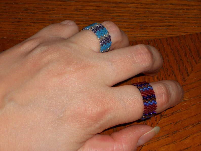 May include: Two beaded rings on a hand. The top ring is blue and silver, the bottom ring is red, blue, and silver.