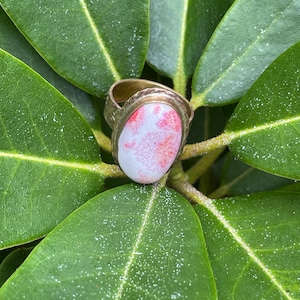Puede incluir: Un anillo de metal con una piedra cabujón de forma ovalada, que presenta una superficie blanca lisa con inclusiones rojas moteadas. La banda del anillo tiene un acabado texturizado y martillado que contrasta con el aspecto pulido de la piedra.