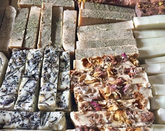 May include: A variety of handmade soap bars with different scents and ingredients. The bars are arranged in rows and feature natural elements like dried flowers, herbs, and seeds.