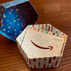 Könnte beinhalten: Eine sechseckige Geschenkbox mit einem blauen und weißen Tupfenmuster. Die Box ist geöffnet und enthält eine weiße Geschenkkarte mit einem roten Amazon-Logo und einem Schneeflockenmuster. Die Geschenkkarte trägt die Aufschrift "amazon" in der oberen rechten Ecke.