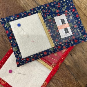 May include: Two rectangular pin cushions with festive holiday patterns. One is navy blue with red and gold stars, the other is red with gold snowflakes. Each has a white felt centre and a clear pocket with sewing supplies.