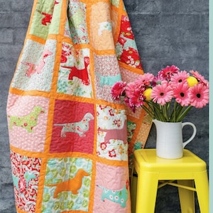 Può includere: Un quilt patchwork con un motivo di cani bassotti in vari colori e motivi. Il quilt è appeso a una scala ed è circondato da un muro di mattoni grigi. Un vaso di fiori rosa è appoggiato su uno sgabello giallo in primo piano.