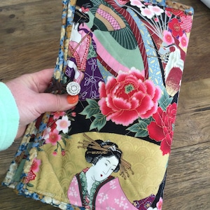 Peut inclure: Une pochette matelassée avec un motif d'inspiration japonaise représentant des geishas, des fleurs et un fond doré. Le tissu est un mélange de noir, d'or, de rose et de rouge.