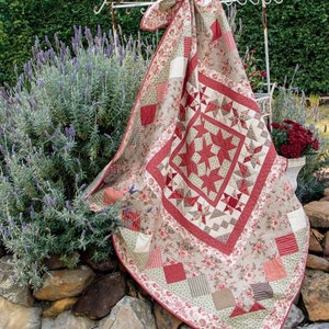 May include: A handmade quilt with a central star pattern in red and cream, surrounded by a floral border. The quilt features various patchwork squares and triangles in coordinating colors. The quilt is draped over a white metal rack outdoors.