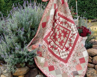 May include: A handmade quilt with a central star pattern in red and cream, surrounded by a floral border. The quilt features various patchwork squares and triangles in coordinating colors. The quilt is draped over a white metal rack outdoors.