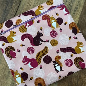 Puede incluir: Una bolsa rosa con cremallera morada, decorada con un estampado repetido de ardillas, bellotas y hojas en tonos burdeos, marrón y blanco. El diseño se repite en la superficie.