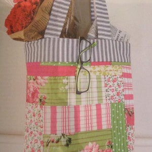 Pode incluir: Uma bolsa tote em patchwork rosa e verde com uma alça listrada. A bolsa está cheia de um buquê de flores vermelhas e um par de óculos.