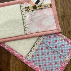 May include: Two handmade fabric sewing organizers. One features a pink floral pattern and a clear pocket with a ruler. The other has a blue gingham pattern with pink polka dots and a ruler. Both have pink trim.