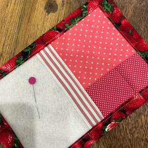 May include: A rectangular fabric needle case featuring a strawberry-patterned border, a white felt panel with a single pink-headed sewing pin, and two sections of pink polka-dot fabric separated by a white and pink striped ribbon trim.
