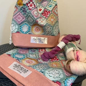 May include: A drawstring project bag and a flat pouch featuring a multi-coloured crochet granny square pattern with a dusty pink polka dot fabric base. Each item has a white label reading Handmade with love by Sues Stitching. Two skeins of yarn are included.