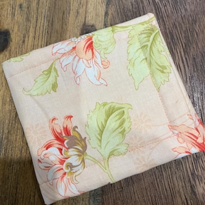 May include: A square fabric coaster featuring a light peach background with a repeating floral pattern of orange and white flowers paired with light green leaves. The coaster has visible stitched edges and a quilted texture throughout the fabric surface.