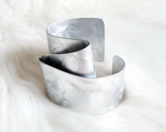 Puede incluir: Un brazalete de metal plateado con un diseño ondulado y texturado. El brazalete está hecho de dos piezas de metal que se unen.