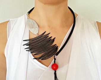 May include: A black and white statement necklace with a large oval pendant and a red bead at the end. The necklace is made of leather and metal.