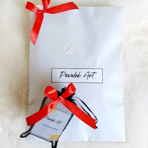May include: A white mailing envelope with a red ribbon tied on top. The envelope has a sticker with the text "Pevalek Art" and a small sticker that says "thank you". Inside the envelope is a small black bag with a red ribbon tied on top. The bag has a white card with the text "Pevalek Art" and a website address.