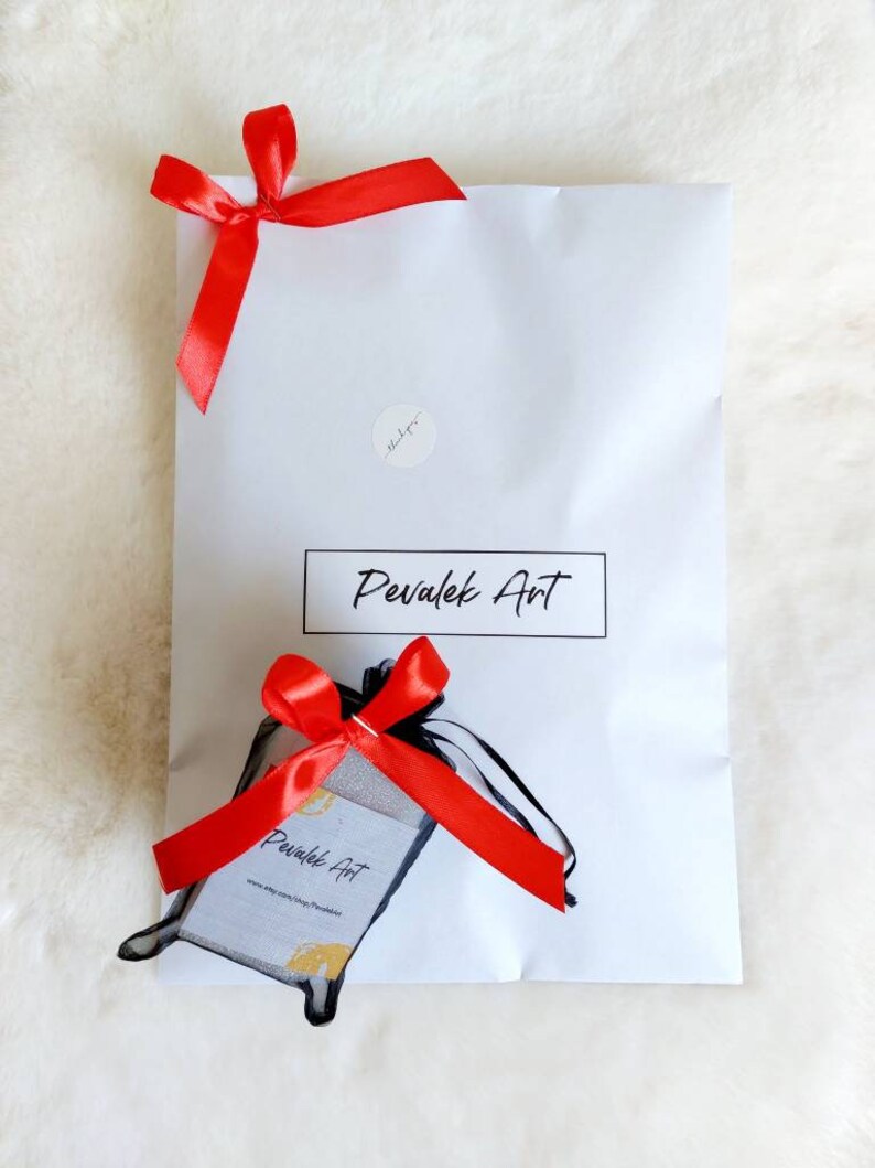 May include: A white mailing envelope with a red ribbon tied on top. The envelope has a sticker with the text "Pevalek Art" and a small sticker that says "thank you". Inside the envelope is a small black bag with a red ribbon tied on top. The bag has a white card with the text "Pevalek Art" and a website address.