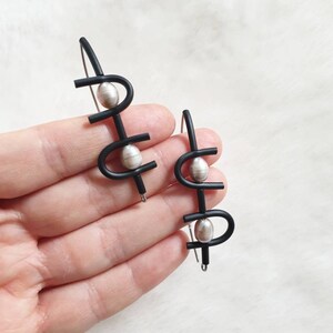 May include: A pair of black, abstract design earrings with silver-colored, oval-shaped beads. The earrings have a modern, minimalist style, with a hook closure. The design features curved and straight lines.
