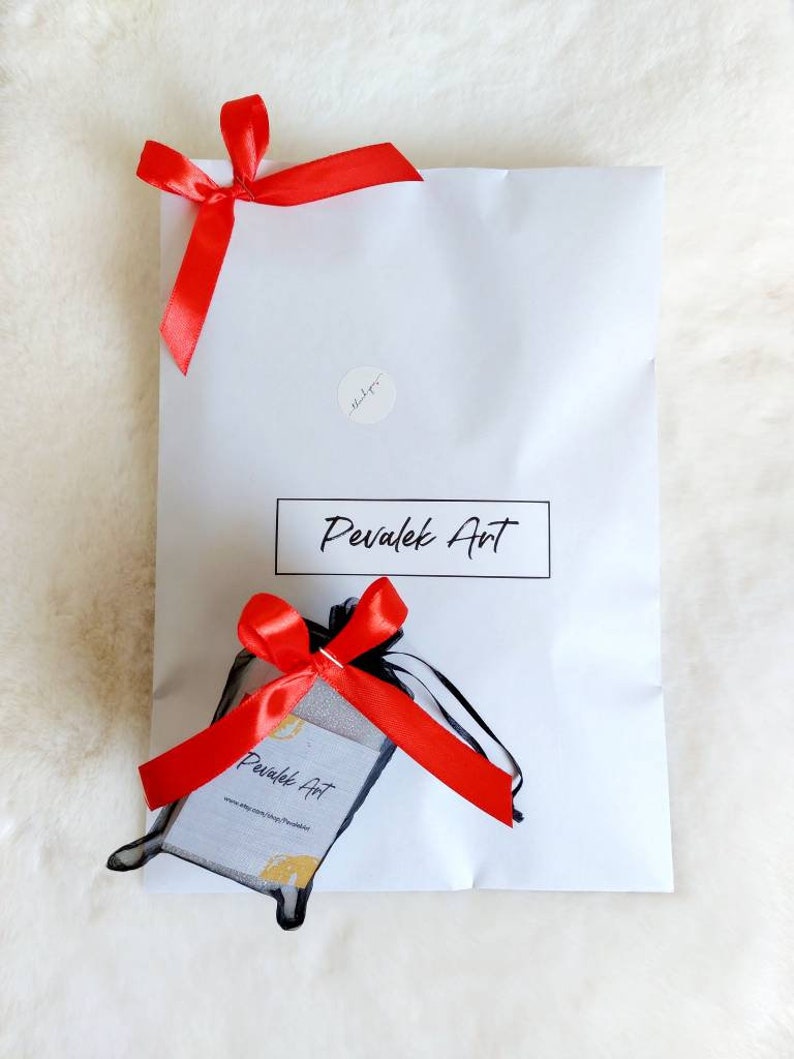 May include: A white paper package tied with a red satin ribbon. The package has the text "Pevalek Art" in a black rectangle. A small black mesh bag with a red ribbon and a business card is also present.