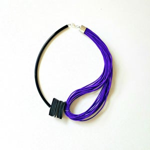 May include: A modern necklace featuring a black, curved band on one side and a bundle of vibrant purple cords on the other. A black, square accent piece adds a bold contrast. The necklace is secured with a silver clasp.