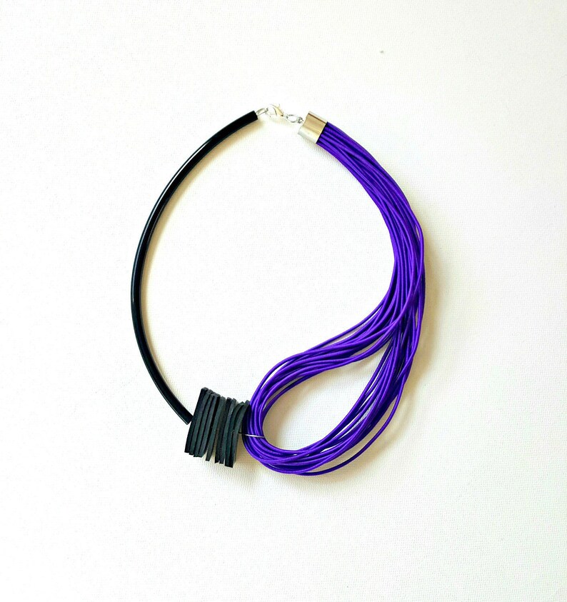 May include: A modern necklace featuring a black, curved band on one side and a bundle of vibrant purple cords on the other. A black, square accent piece adds a bold contrast. The necklace is secured with a silver clasp.