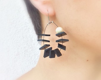 May include: A modern, geometric earring featuring a black rectangular design with a small, off-white sphere. The earring is suspended from a silver-tone wire. The design is abstract and minimalist, suitable for everyday wear.