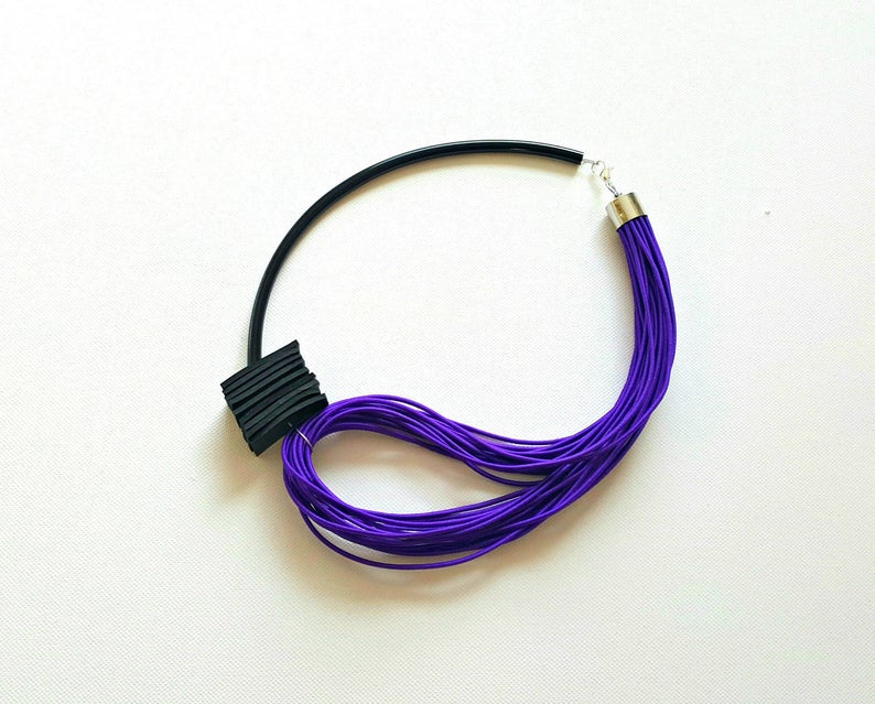 May include: A modern necklace featuring a black, curved band and a cluster of vibrant purple cords. A black, textured square detail is attached to the band. The necklace is secured with a silver clasp, creating a striking contrast.