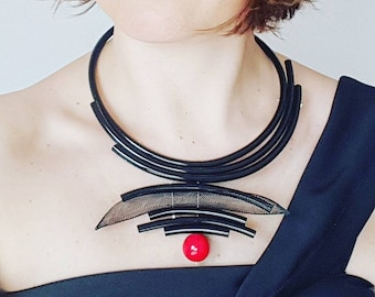 Puede incluir: Un collar gargantilla negro con un diseño geométrico y un colgante de cuentas rojas.