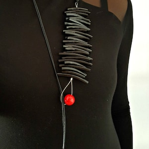 Pode incluir: Um colar preto com um longo pingente feito de tubos pretos empilhados e uma única conta vermelha. O colar é usado por uma pessoa que está vestindo uma camisa preta.