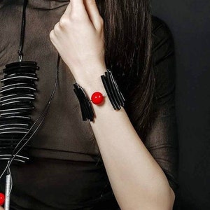 Op de afbeelding: Een zwarte armband met een rode kraal in het midden. De armband is gemaakt van dunne, zwarte stroken materiaal die in een geometrisch patroon zijn gerangschikt.
