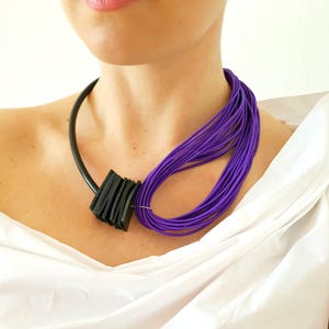 May include: A modern necklace featuring a black, geometric pendant and a cluster of vibrant purple cords. The necklace has a black, curved band that rests around the neck. The purple cords are looped to the side, creating a bold, artistic statement.