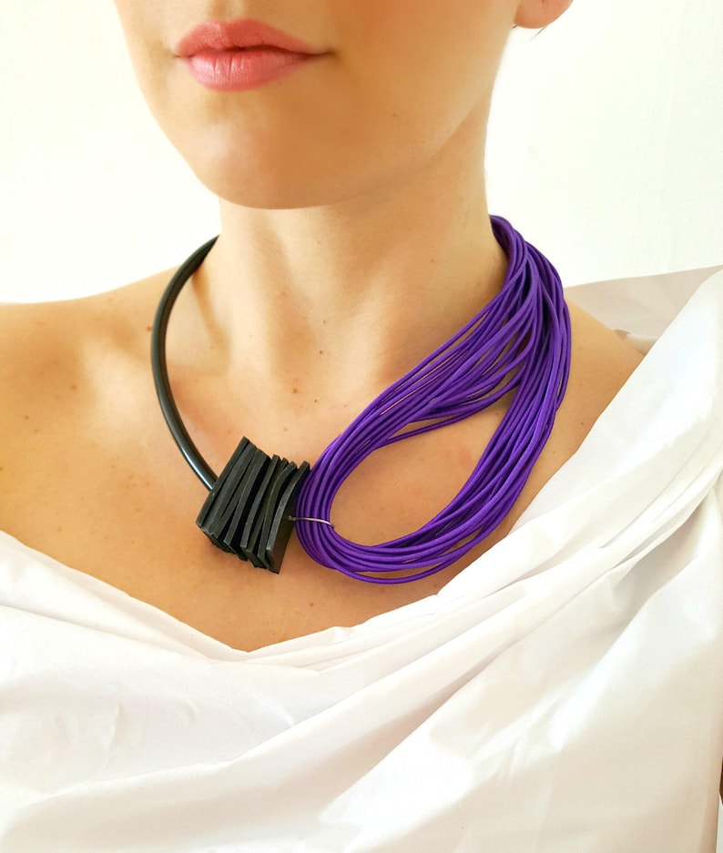 May include: A modern necklace featuring a black, geometric pendant and a cluster of vibrant purple cords. The necklace has a black, curved band that rests around the neck. The purple cords are looped to the side, creating a bold, artistic statement.