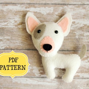 Pu&ograve; includere: Un cane di razza bull terrier in feltro bianco con orecchie rosa, occhi neri e naso marrone. Il cane &egrave; in piedi su una superficie di legno marrone chiaro. Il testo "PDF PATTERN" &egrave; su un'etichetta gialla.