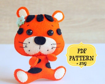 May include: A felt tiger plush toy with black stripes and a small flower on its head. The toy is sitting on a white surface with a yellow sign that says "PDF PATTERN + SVG".