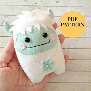 May include: PDF pattern for a felt Yeti plush toy. The Yeti is white with a green face, black eyes, and a pink blush. It is wearing a white flower with a blue centre.