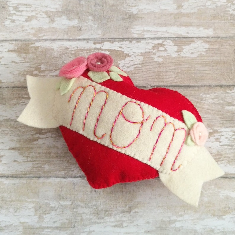 May include: A red felt heart with a white felt banner that says "MOM" in pink embroidery. The heart is decorated with pink felt flowers and green felt leaves.