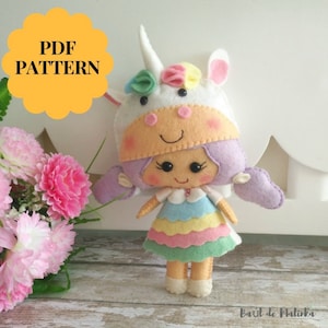 May include: A felt doll wearing a unicorn costume. The doll has purple hair, a rainbow dress, and a white unicorn head with a pink horn. The doll is standing on a wooden surface. PDF PATTERN.