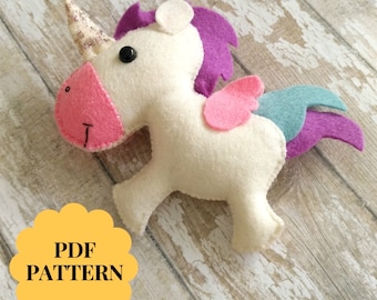 May include: A white felt unicorn with a pink nose, purple mane, and teal and purple tail. The unicorn has a glittery horn and pink wings. The image includes the text "PDF PATTERN".