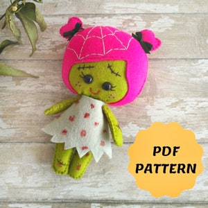 May include: A felt doll with a pink spiderweb hat and a white dress with red polka dots. The doll has green skin and a stitched-on smile. The text "PDF PATTERN" is on a yellow circle in the background.