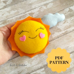 May include: A felt sun with a smiling face and pink hearts for cheeks. The sun has a light blue cloud next to it. The text "PDF PATTERN" is on a yellow circle in the lower right corner of the image.
