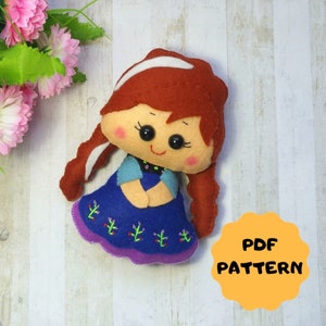 May include: A felt doll pattern for a girl with long red hair, wearing a blue dress with purple trim and green embroidery. The pattern is available as a PDF download. The text "PDF PATTERN" is on a yellow circle.