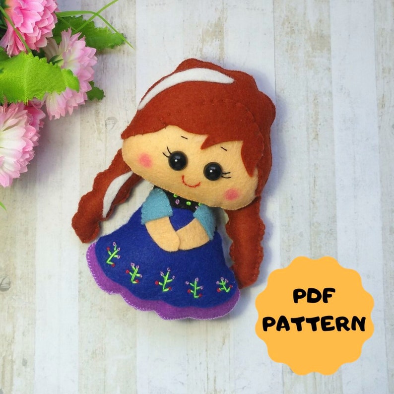 May include: A felt doll pattern for a girl with long red hair, wearing a blue dress with purple trim and green embroidery. The pattern is available as a PDF download. The text "PDF PATTERN" is on a yellow circle.