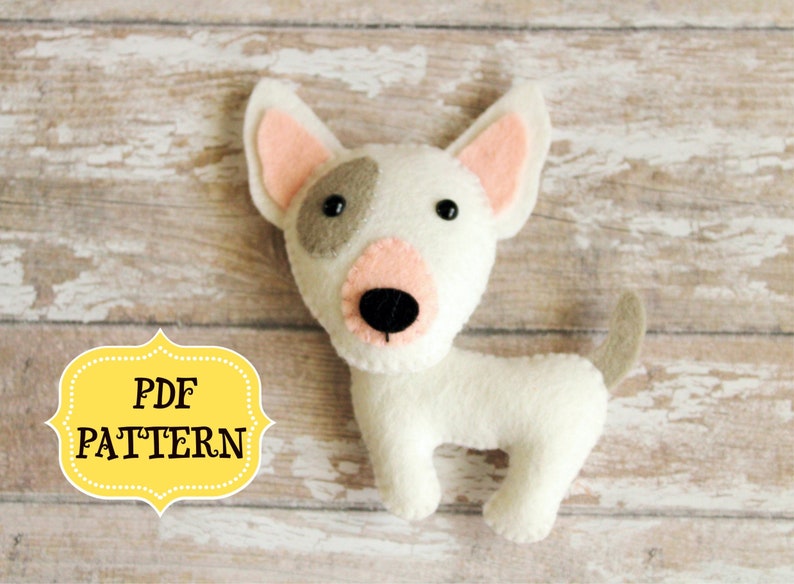 Pu&ograve; includere: Un cane di razza bull terrier in feltro bianco con orecchie rosa, occhi neri e naso marrone. Il cane &egrave; in piedi su una superficie di legno marrone chiaro. Il testo "PDF PATTERN" &egrave; su un'etichetta gialla.