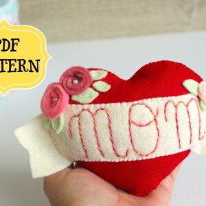 May include: A red felt heart with a white felt banner that says "mom" embroidered in pink and yellow thread. The heart is decorated with three pink felt flowers with white pearl centers and green felt leaves.