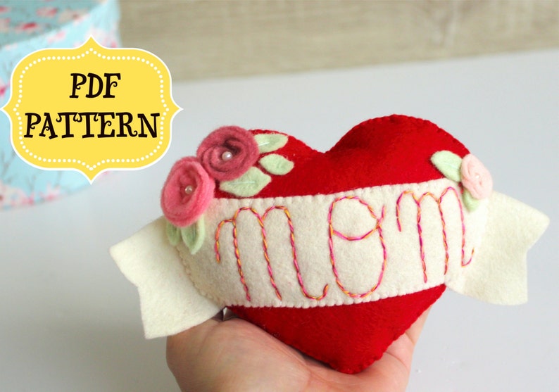 May include: A red felt heart with a white felt banner that says "mom" embroidered in pink and yellow thread. The heart is decorated with three pink felt flowers with white pearl centers and green felt leaves.
