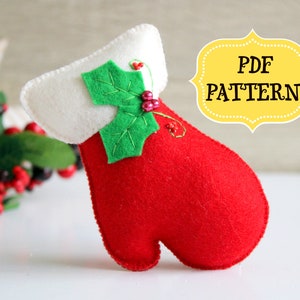 May include: A red felt mitten with a white felt cuff and a green felt holly leaf with red berries. The mitten is a sewing pattern for a Christmas ornament. The text "PDF PATTERN" is on a yellow label.