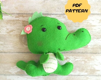 May include: A green felt stuffed toy crocodile with a pink flower on its head. The crocodile has black eyes and a white belly. The toy is sitting on a wooden surface. PDF PATTERN.